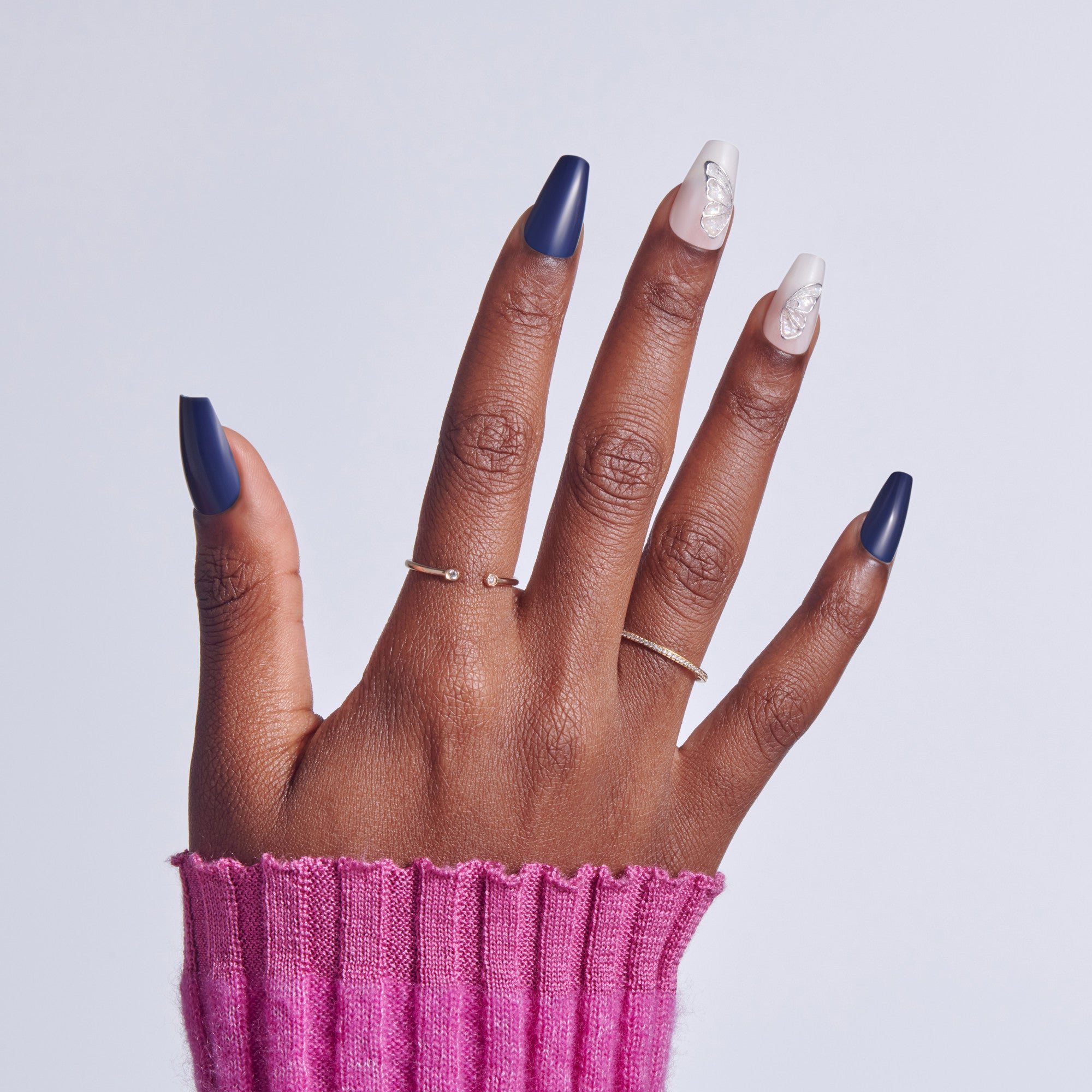 Hand with navy and white nail polish, wearing delicate rings, against pink sleeve.