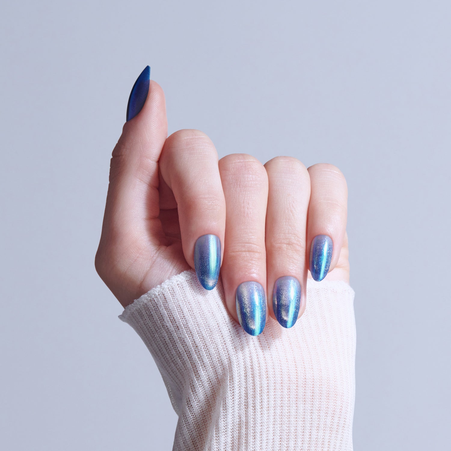 Hand with iridescent blue nail polish on almond-shaped nails, wearing white sleeve.
