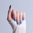 Hand with iridescent blue nail polish on almond-shaped nails, wearing white sleeve.