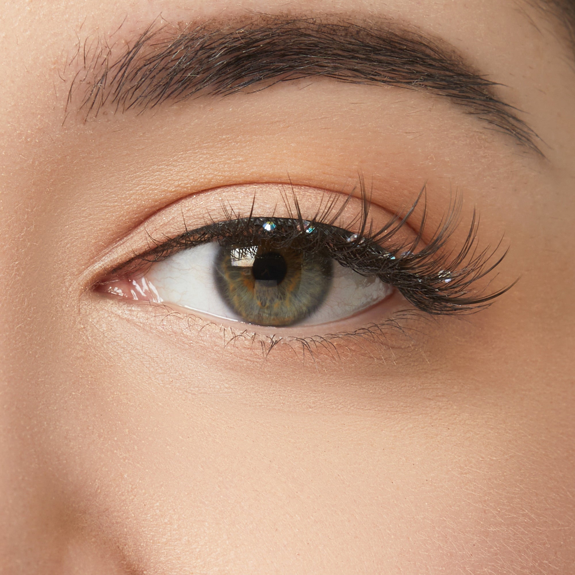 The image is a close-up of a human eye. The eye has a greenish-brown iris with a dark outer ring. The eyelashes are long and well-defined, and the eyebrow above is neatly groomed. The skin around the eye is smooth, with a natural, light complexion. The eyelid has a subtle, neutral-toned eyeshadow applied, enhancing the eye&