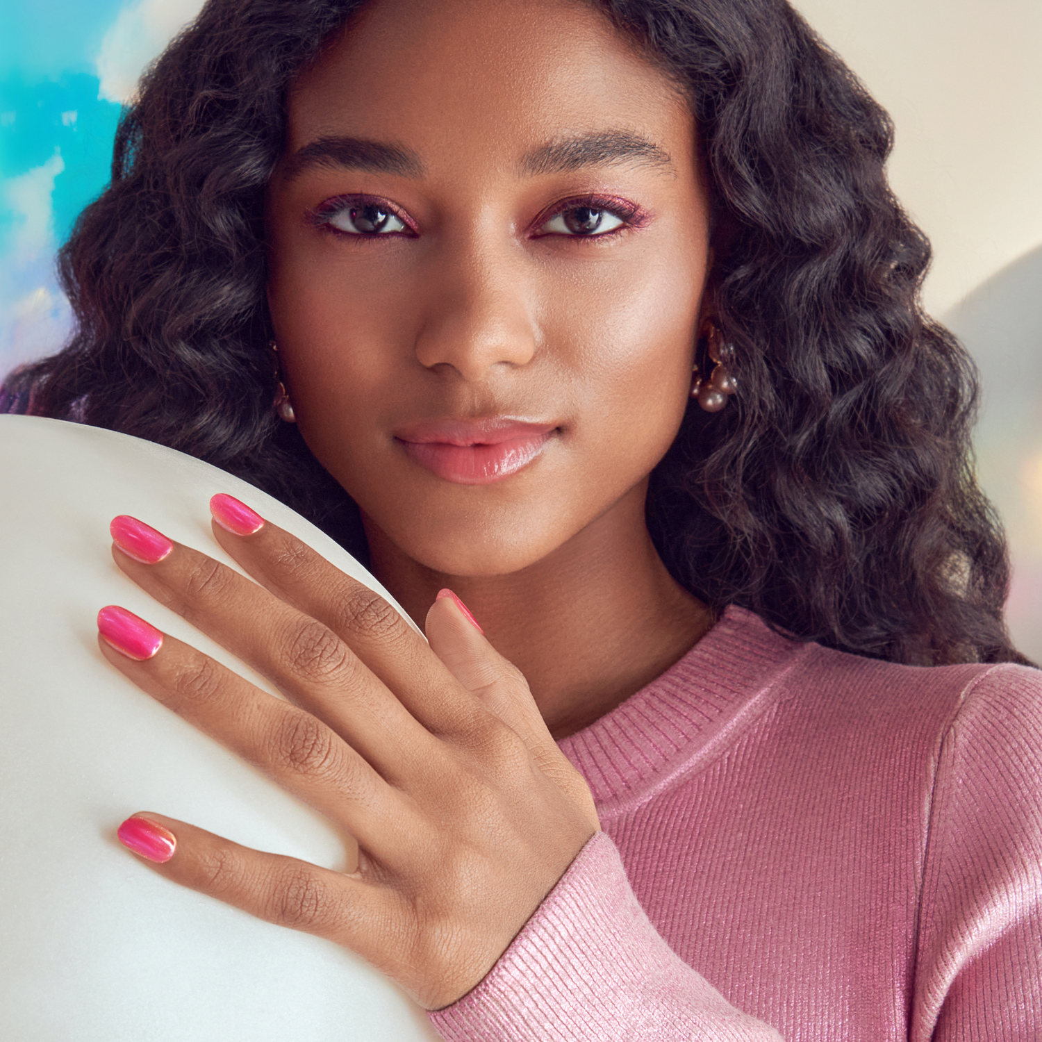 The image features a young woman with long, wavy hair and a warm complexion. She is looking directly at the camera with a soft expression. Her makeup includes subtle pink tones, enhancing her natural beauty. She is wearing a light pink sweater and has well-manicured nails painted in a matching shade of pink. The background is softly blurred, suggesting a bright and airy atmosphere. The overall vibe is fresh and inviting.