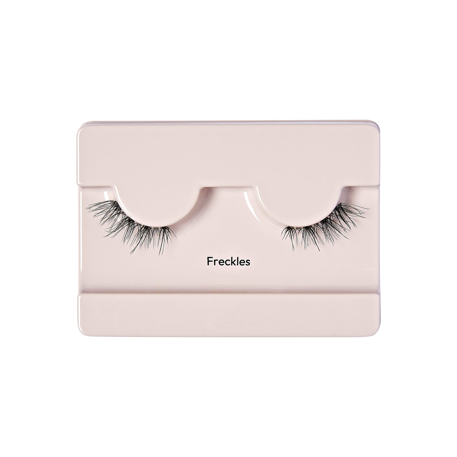 The image shows a pair of false eyelashes displayed in a light pink or beige plastic case. The case has two curved indentations where the eyelashes are placed, mimicking the shape of an eye. The eyelashes are long and wispy, with a natural look. Below the indentations, the word "Freckles" is printed in black text. The overall presentation is clean and minimalistic.