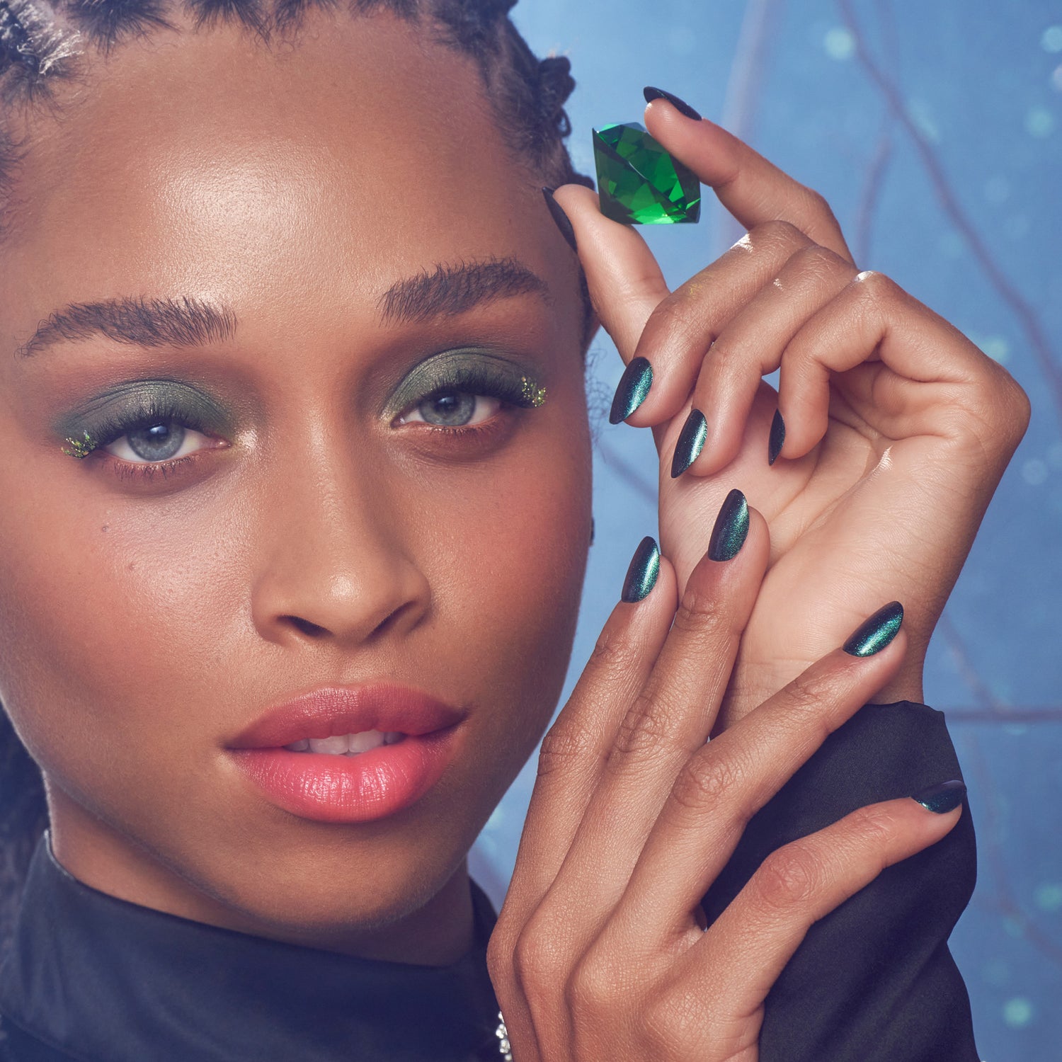 The image features a close-up of a person with striking makeup and a captivating expression. They have dark, braided hair and are holding a green gemstone delicately between their fingers. Their eyes are accentuated with bold green eyeshadow, and their nails are painted in a glossy dark shade that complements the overall look. The background has a soft, ethereal quality, enhancing the vibrant colors and the subject&