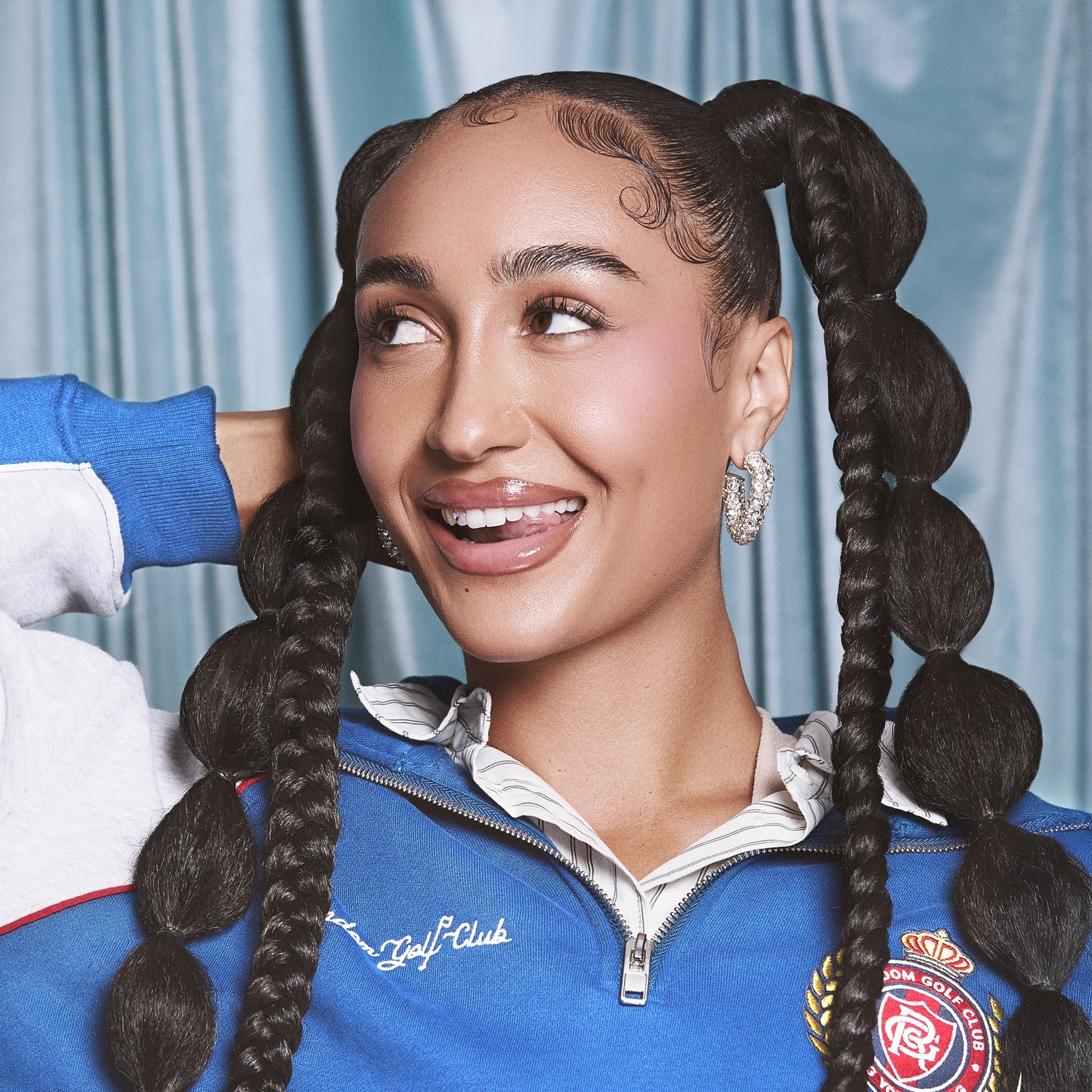Woman with glossy makeup and styled baby hairs, wearing sparkling hoop earrings and a blue zip-up jacket against a light blue curtain background.