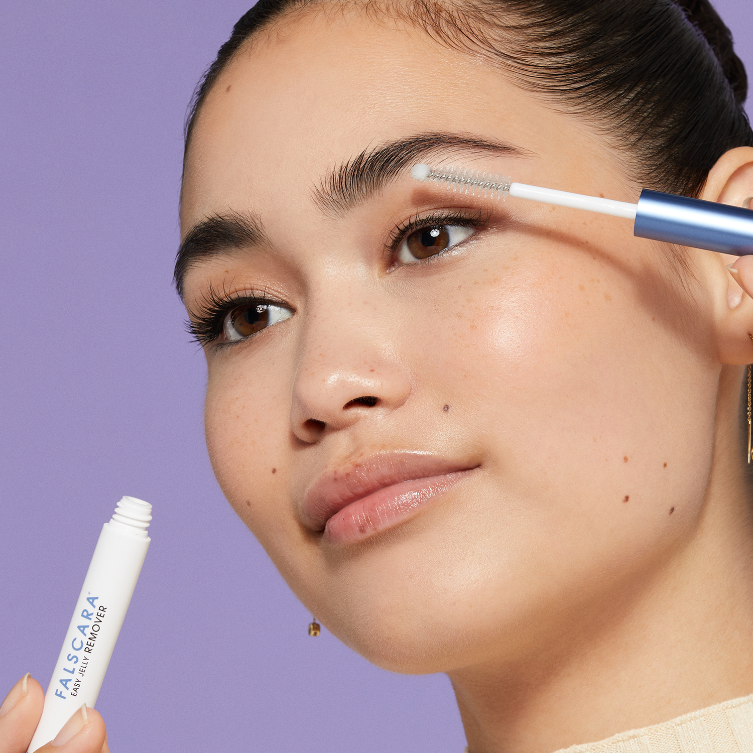 Close-up of a woman applying a clear gel eyelash and eyebrow brush from a white tube labeled \