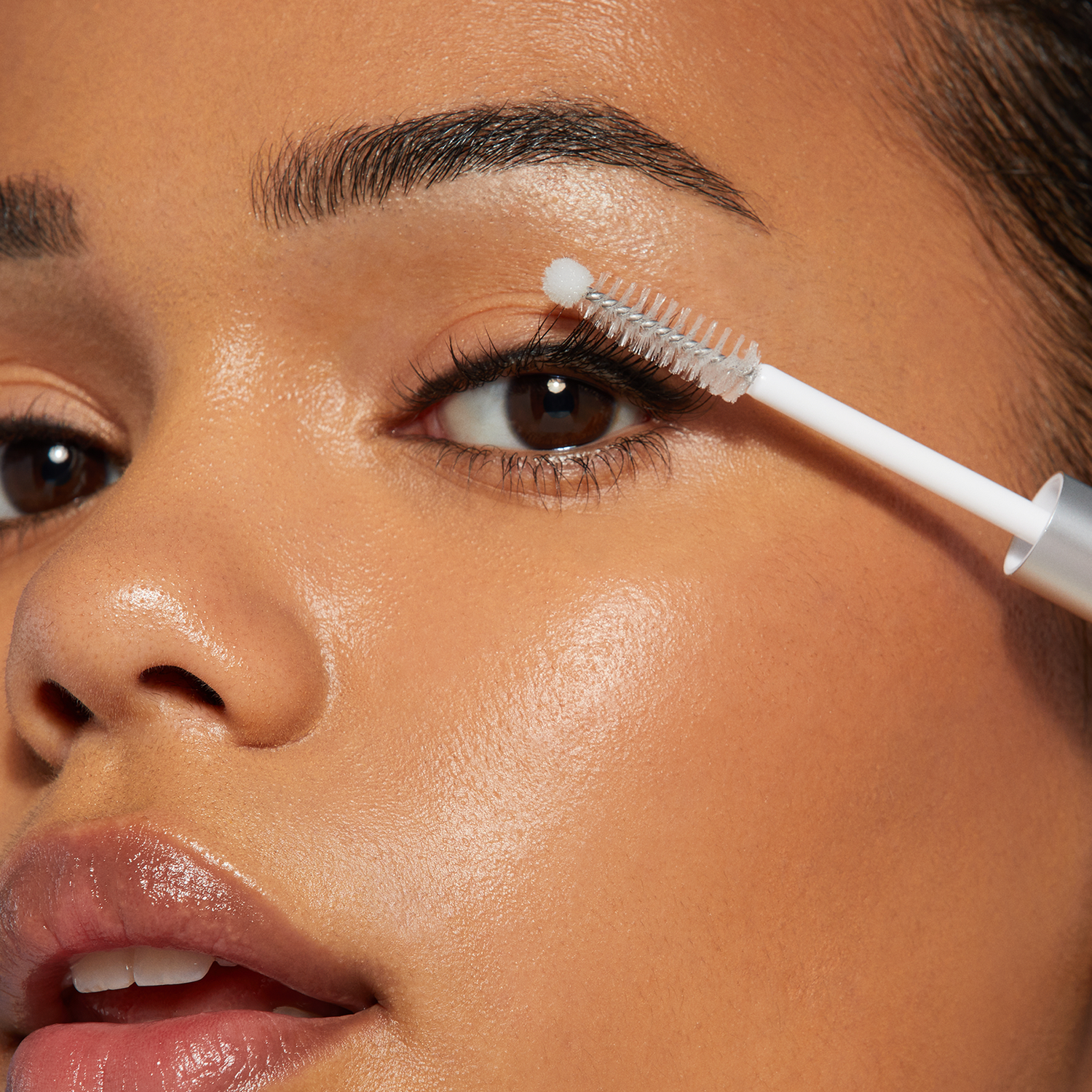 Close-up of a brown-eyed person applying clear lash adhesive with a white applicator wand featuring a small round tip near the upper eyelashes.