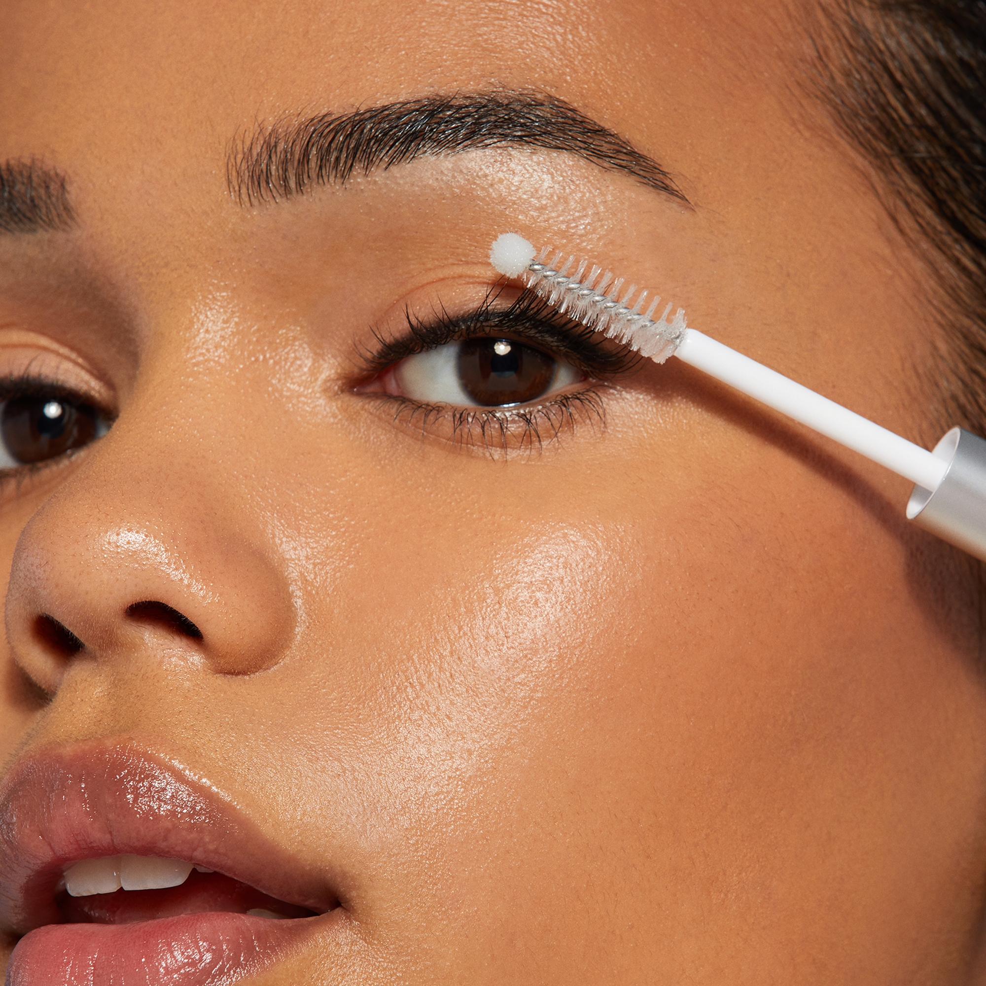 Close-up of a person applying clear lash bonding gel near their upper eyelashes with a white applicator brush against smooth skin.