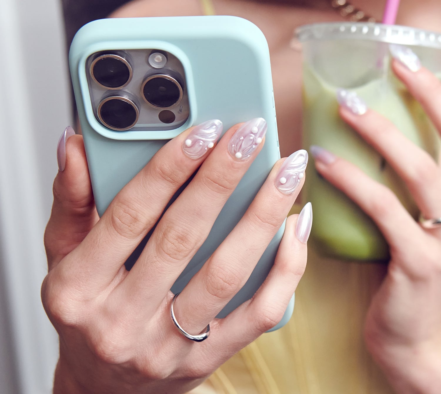 Person holding a phone with a light blue case and a drink, wearing a ring.