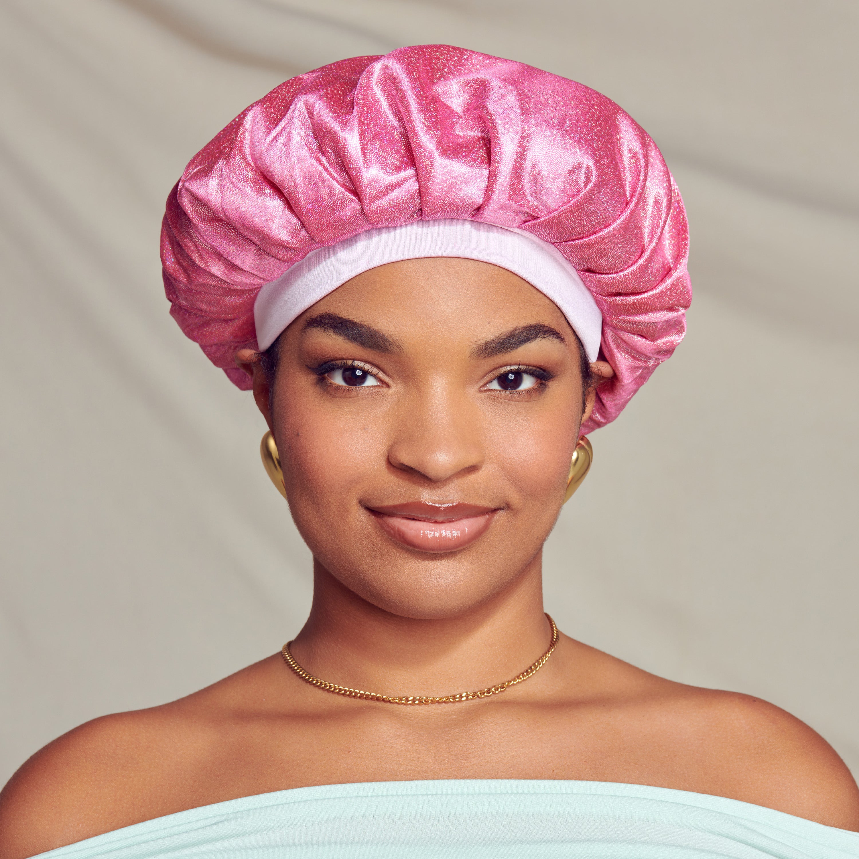 Young woman wearing a shiny pink glitter bonnet cap with a light pink band, gold earrings, and a thin gold chain necklace against a neutral background.
