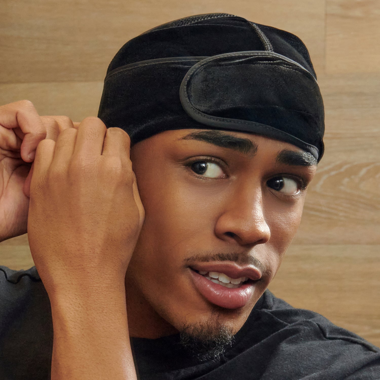 Young man wearing a black velvet durag, shown adjusting the strap over his ear against a light wood background.