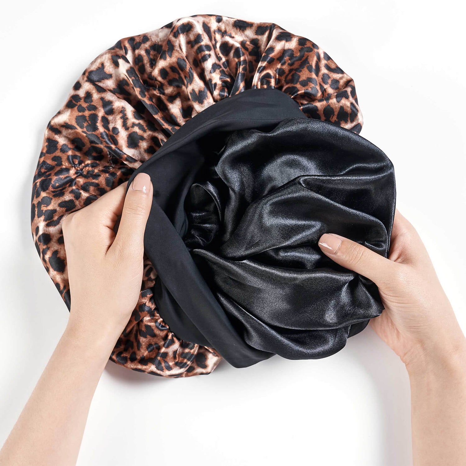 Two hands holding open a satin sleep cap with a leopard print exterior and a black satin interior, shown against a plain white background.