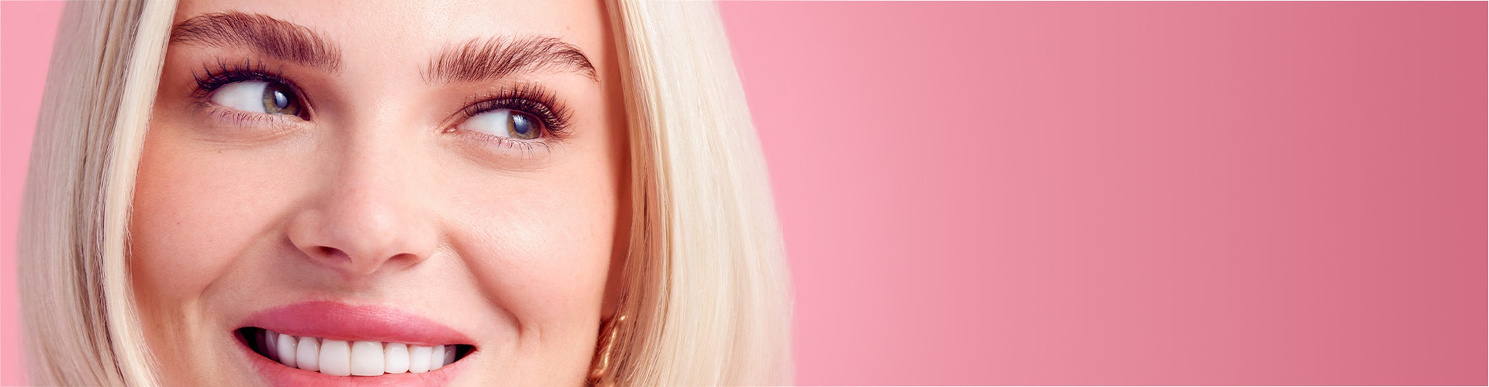 Woman with blonde hair smiling, wearing KISS lashes against pink background