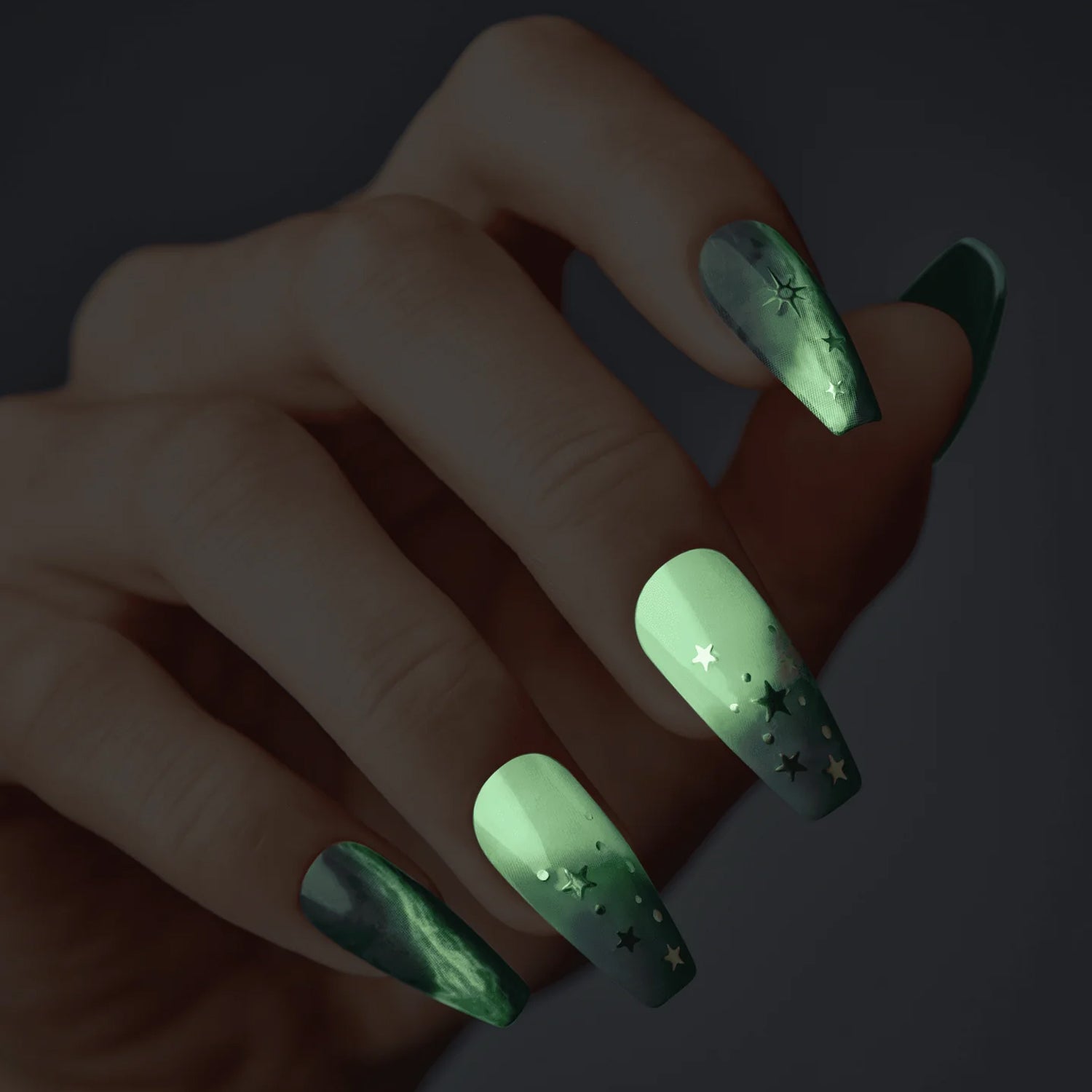 Hand with long, coffin-shaped press-on nails in a green ombre design featuring shiny star and dot accents, shown against a dark background.