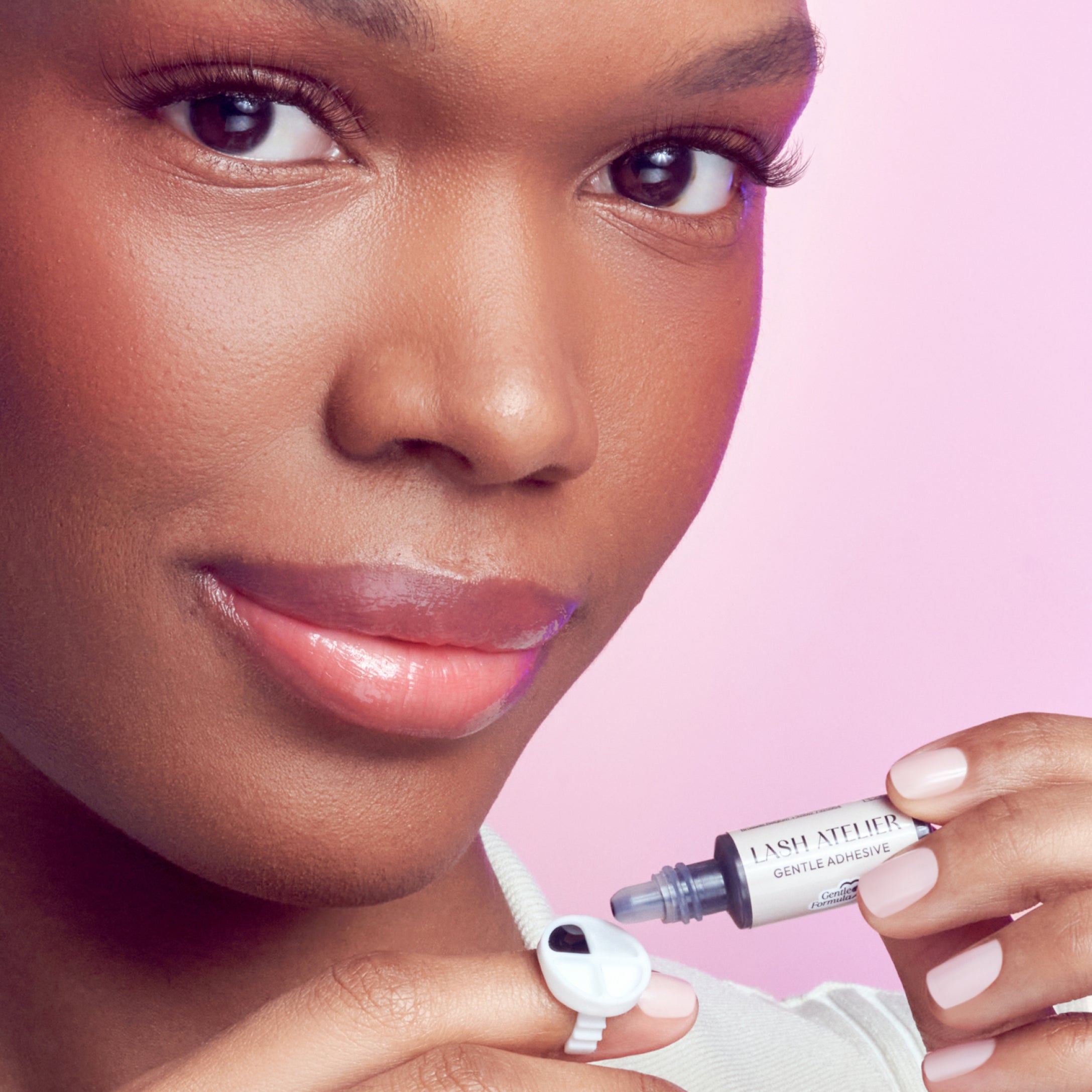 Woman holding Lash Attire Gentle Adhesive lash glue against pink background.