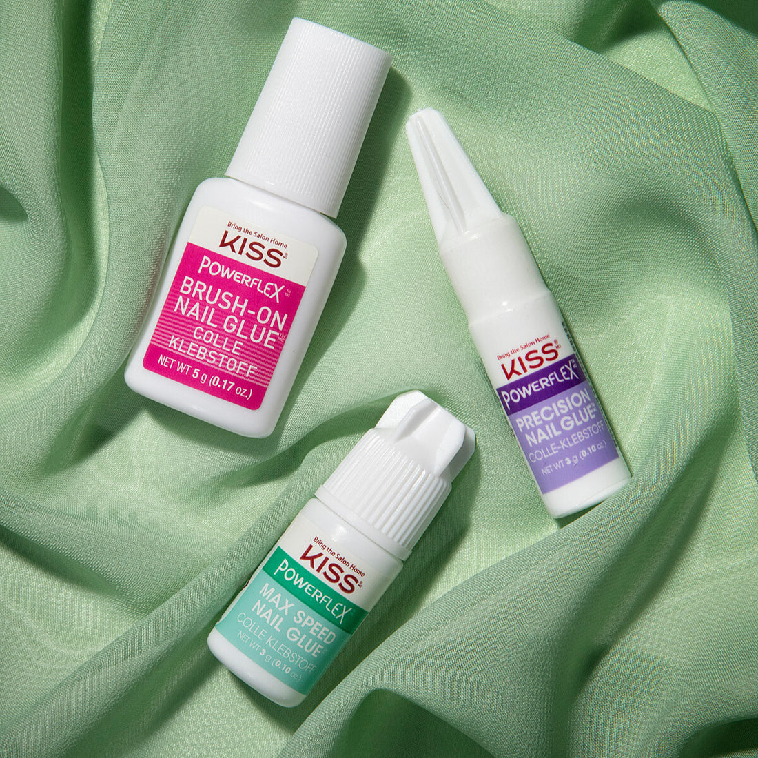 Three small white bottles of KISS Powerflex nail glues—Brush-On, Precision, and Max Speed—lay on soft light green fabric.