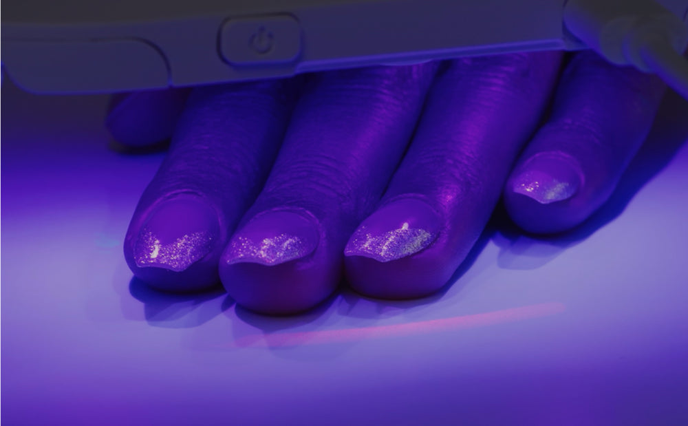 Nail tips with glittery purple polish under UV light during application demonstration.