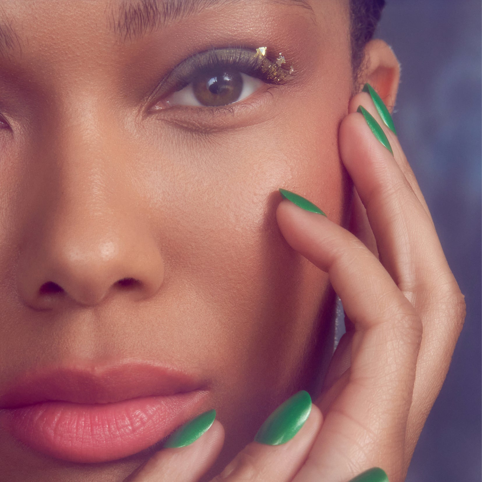 Woman with gold eyeshadow and green nail art touching face against purple background.