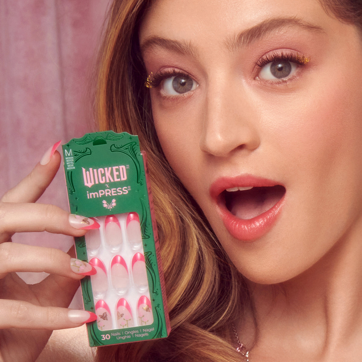 Woman holding Wicked imPRESS press-on nails with pink and white design against pink background.
