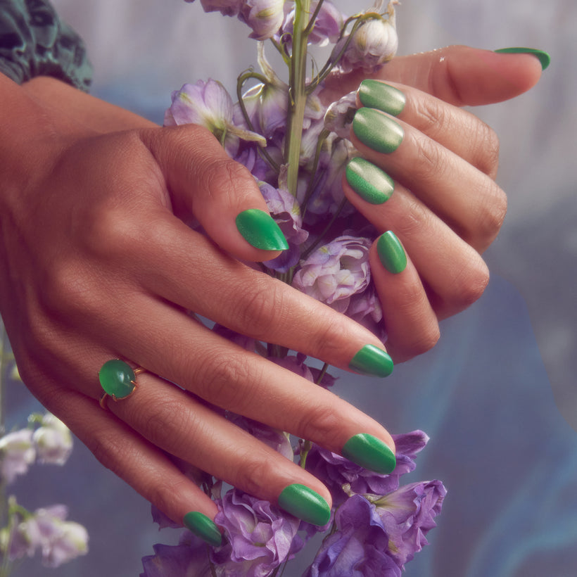 Green manicured nails with shimmer finish holding purple flowers, gold ring visible.