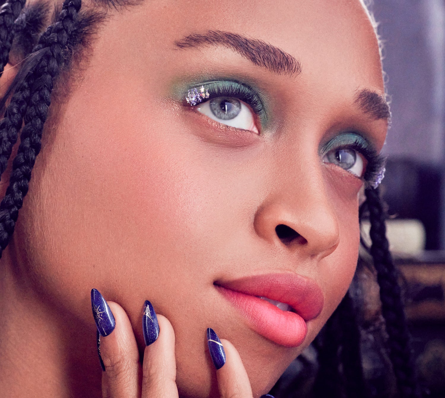Woman with teal eyeshadow, rhinestone accents, braided hair, and navy stiletto nails.