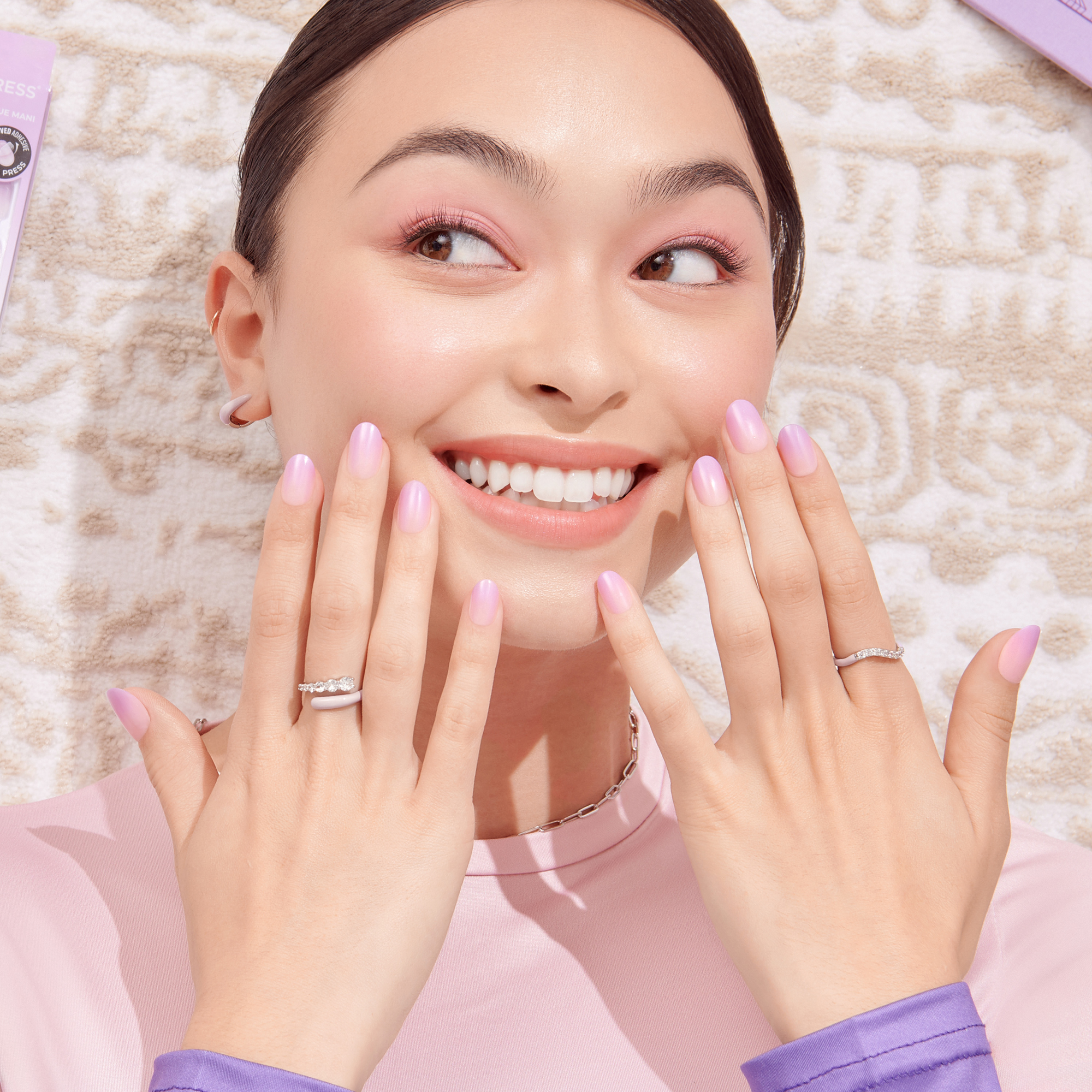 The image features a young woman with a joyful expression, showcasing her manicured hands. She has light pink nail polish on her nails and is wearing multiple rings on her fingers. Her makeup includes soft pink tones, and her hair is styled neatly. The background appears to have a light, textured surface, possibly a rug or fabric, with some beauty products visible nearby, contributing to a fresh and vibrant aesthetic.
