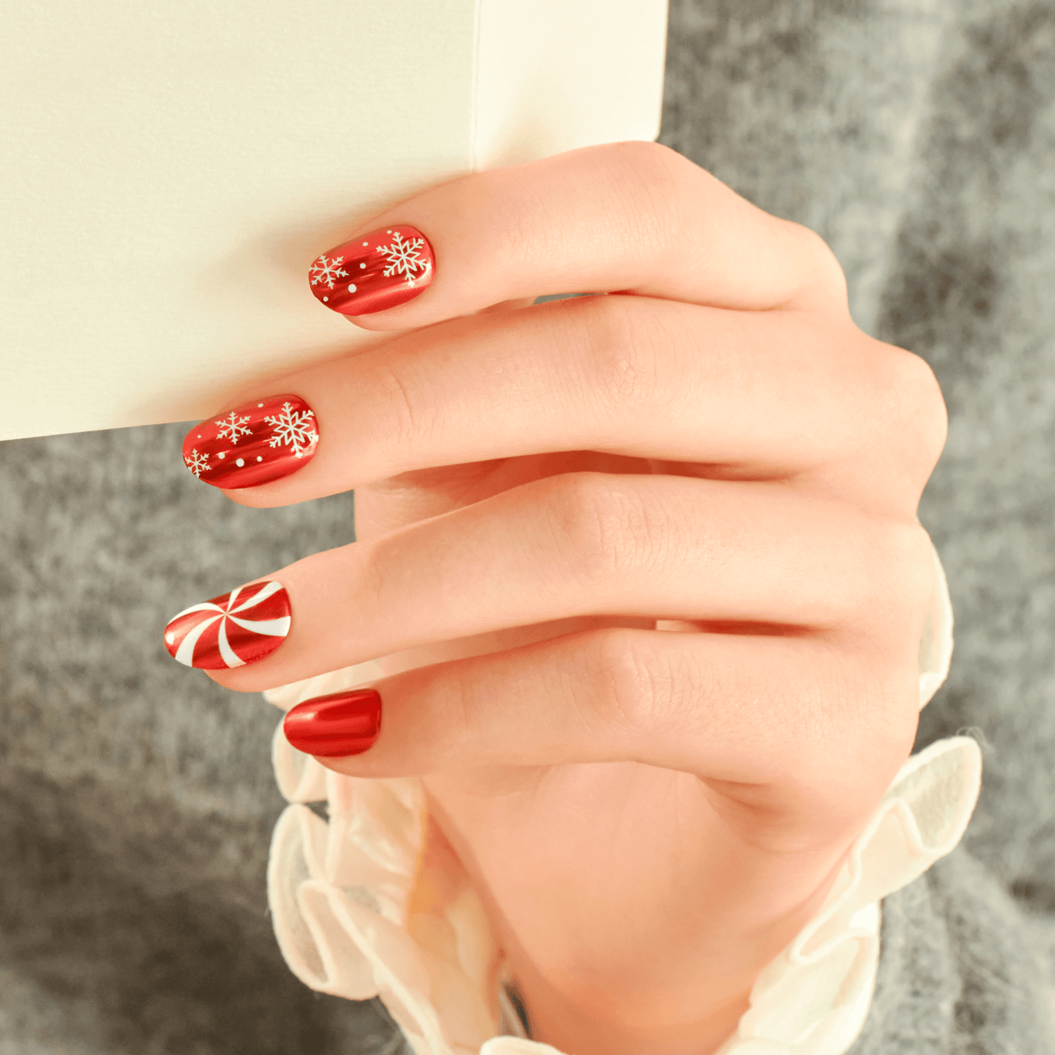 Red festive nails with snowflakes and candy cane designs on hand against gray background.