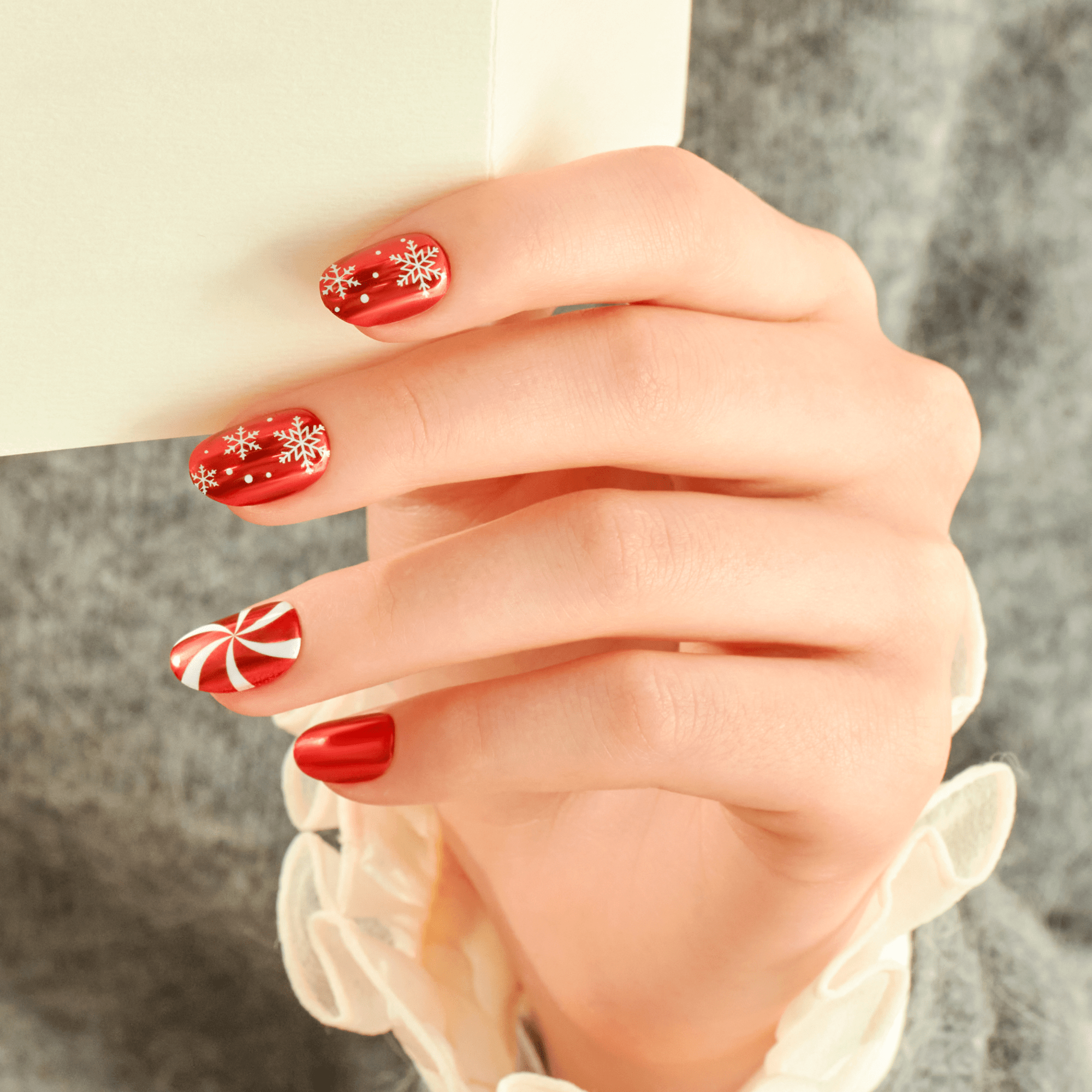 Red festive nails with snowflakes and candy cane designs on hand against gray background.