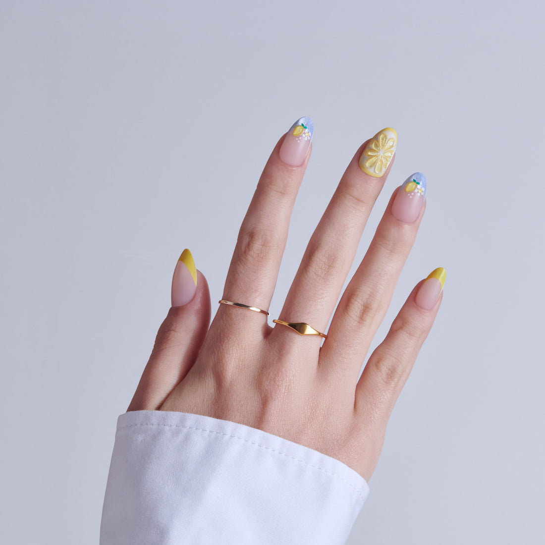 Hand with lemon-themed nail art featuring yellow accents and blue tips, wearing gold rings.