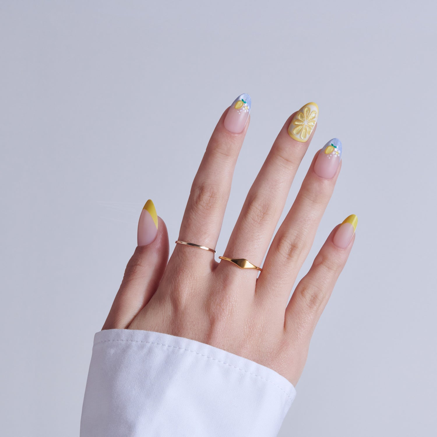 Hand with lemon-themed nail art featuring yellow accents and blue tips, wearing gold rings.