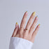 Hand with lemon-themed nail art featuring yellow accents and blue tips, wearing gold rings.