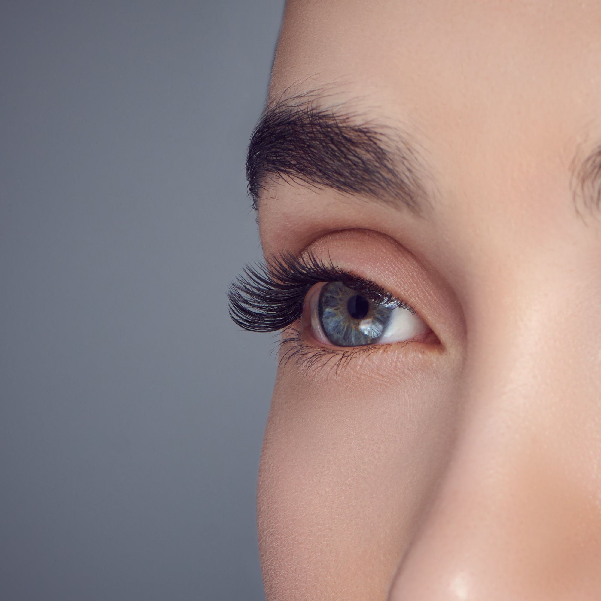Close-up of a blue eye with dense, curled, black false eyelashes applied on the upper lid, showing natural skin and eyebrow against a neutral gray background.