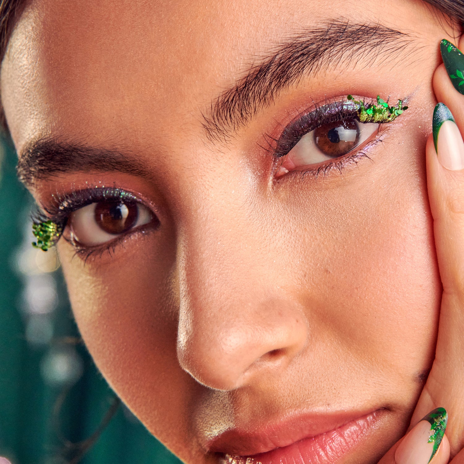 Close-up of eye makeup with green glitter eyeshadow, burgundy liner, and matching green nail art.