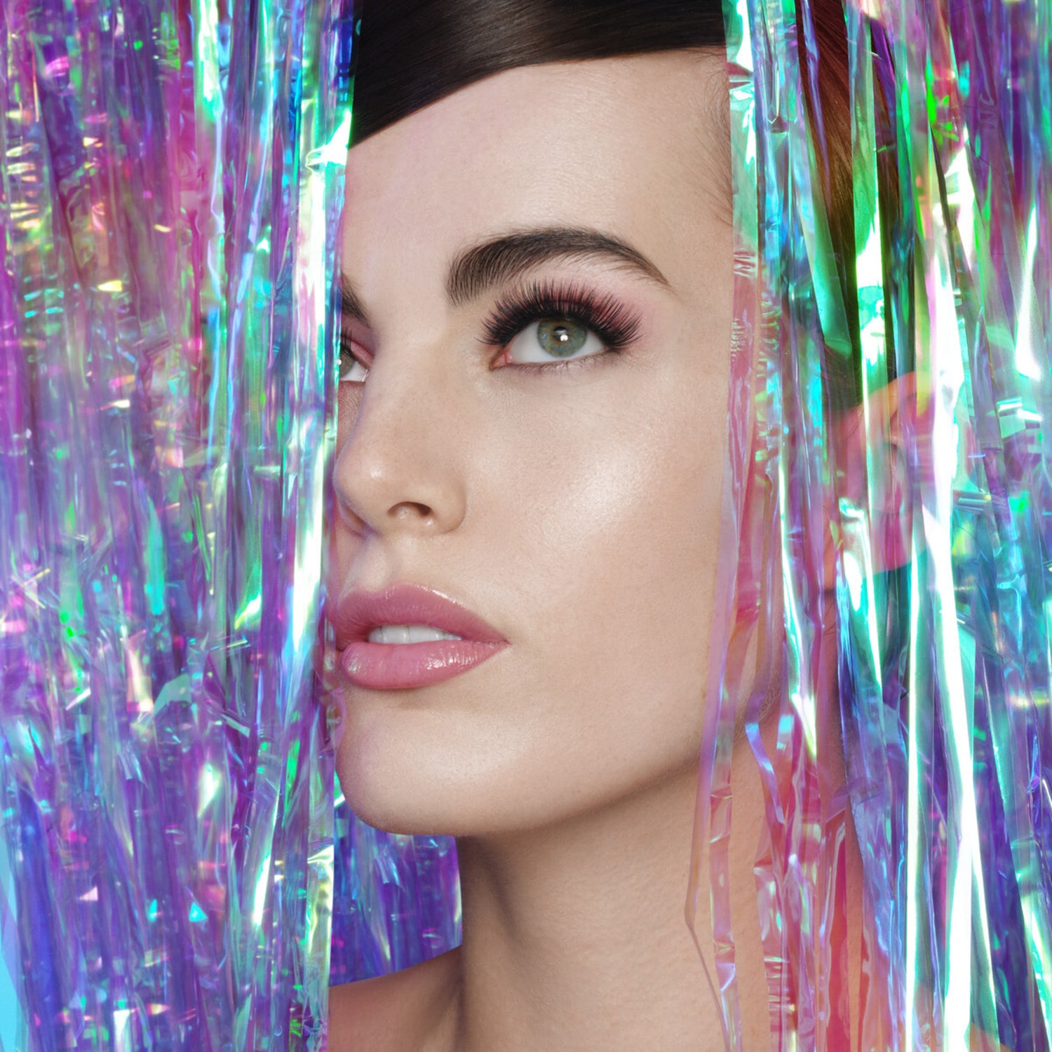 Close-up of a woman with fair skin and green eyes wearing dramatic, long false eyelashes, framed by iridescent purple and blue tinsel strands.