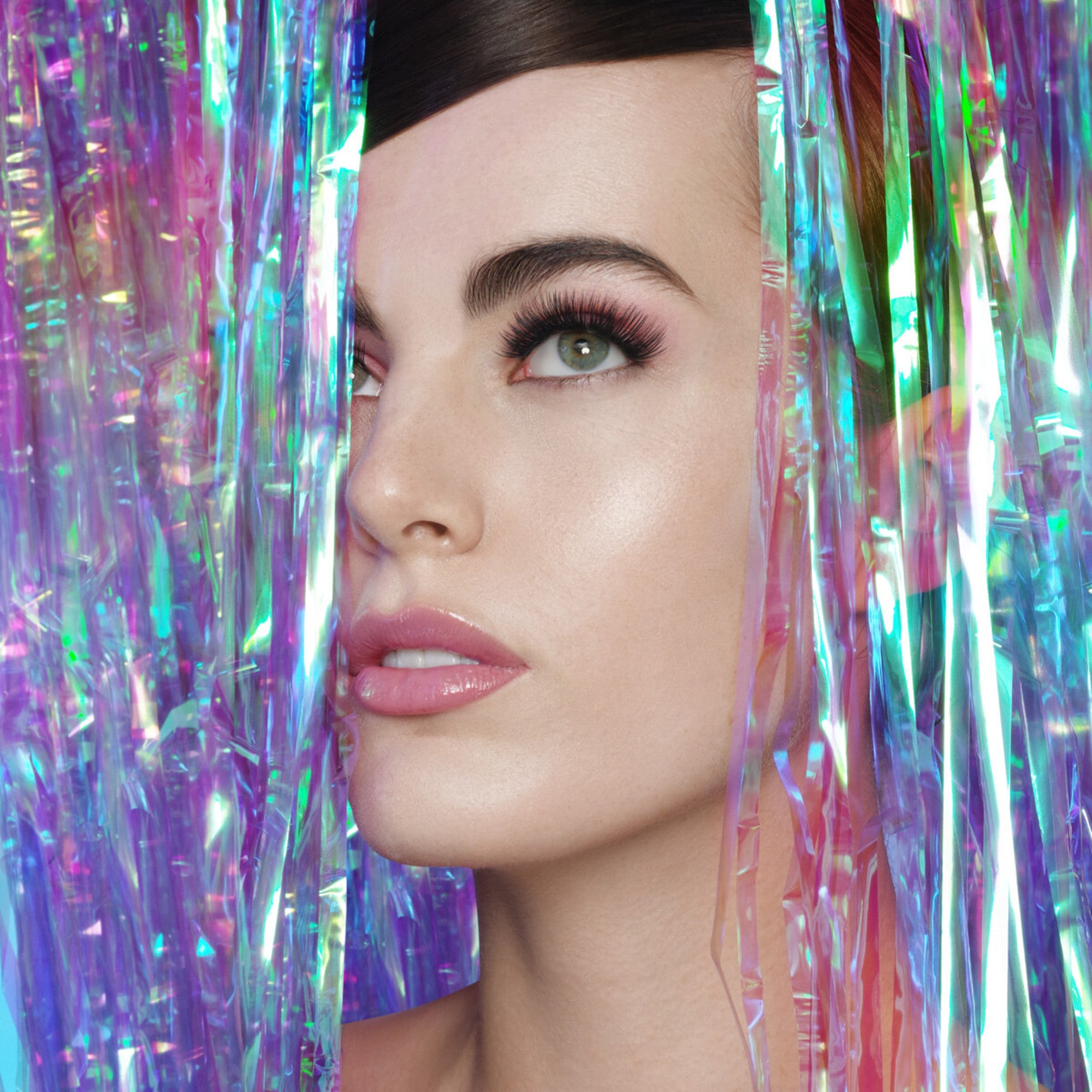 Close-up of a woman with fair skin and green eyes wearing dramatic, long false eyelashes, framed by iridescent purple and blue tinsel strands.