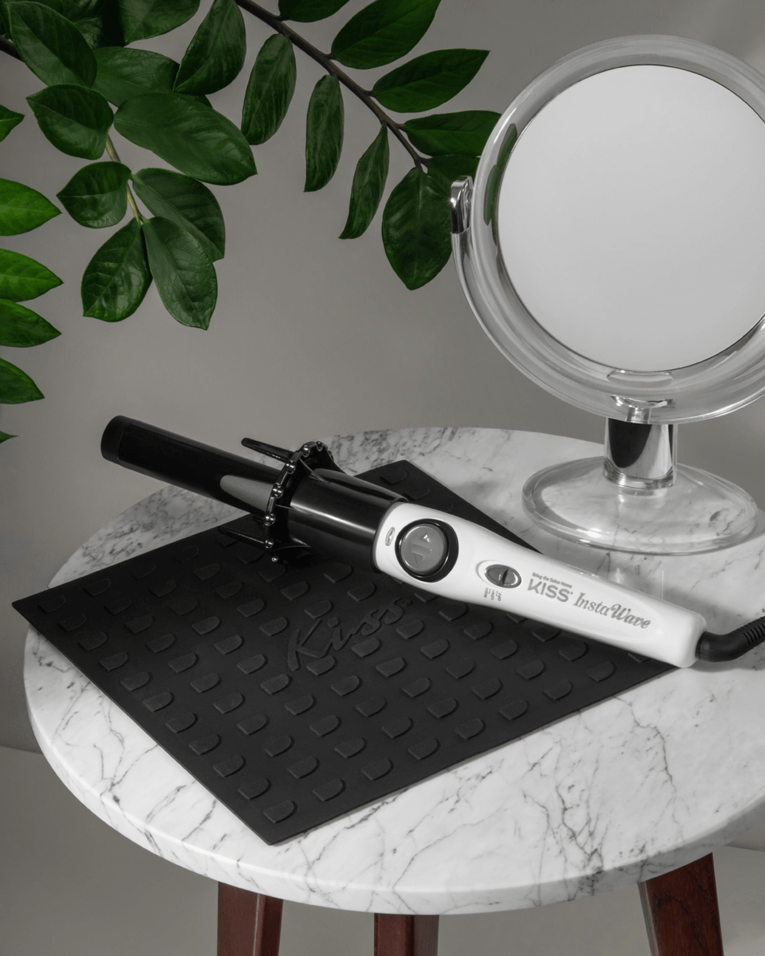 White and black Instawave automatic hair curler resting on a black Kiss-branded heat-resistant mat on a round marble table, next to a round makeup mirror and green leaves.