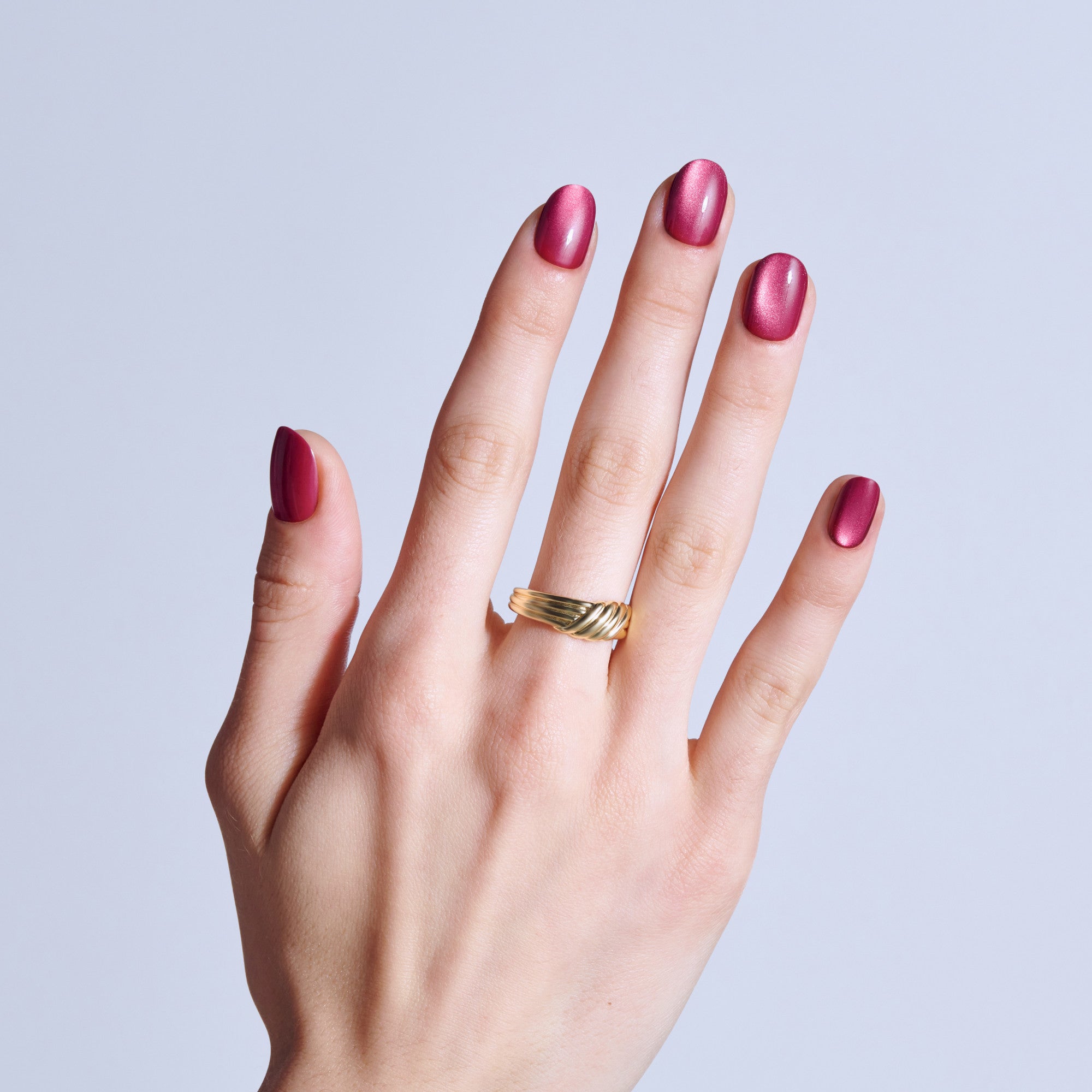 Hand with metallic pink manicure and gold twisted ring on light blue background.