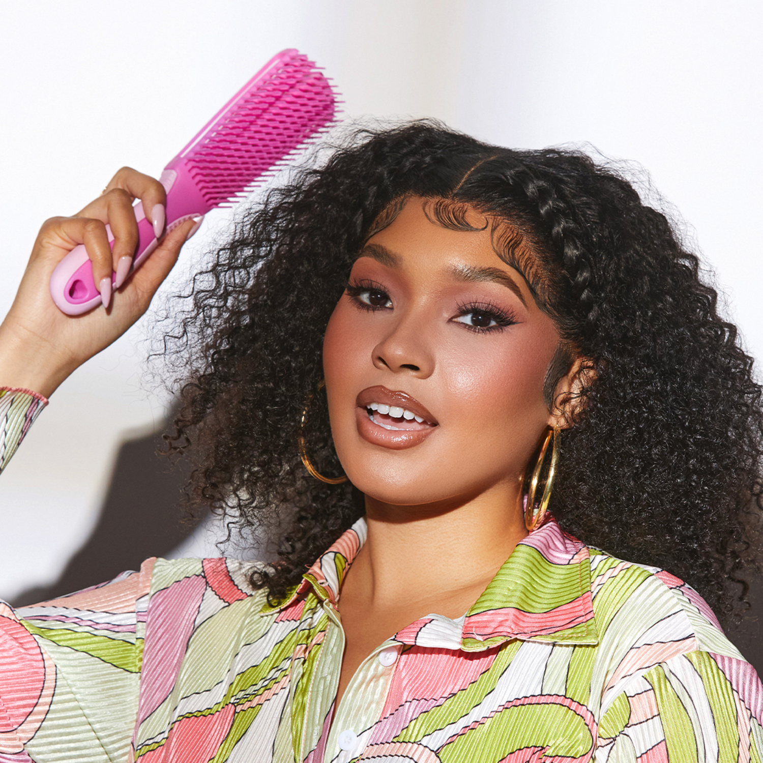 Woman with curly hair and braided front sections holds a pink non-slip detangling brush above her head, wearing a colorful patterned shirt and gold hoop earrings.