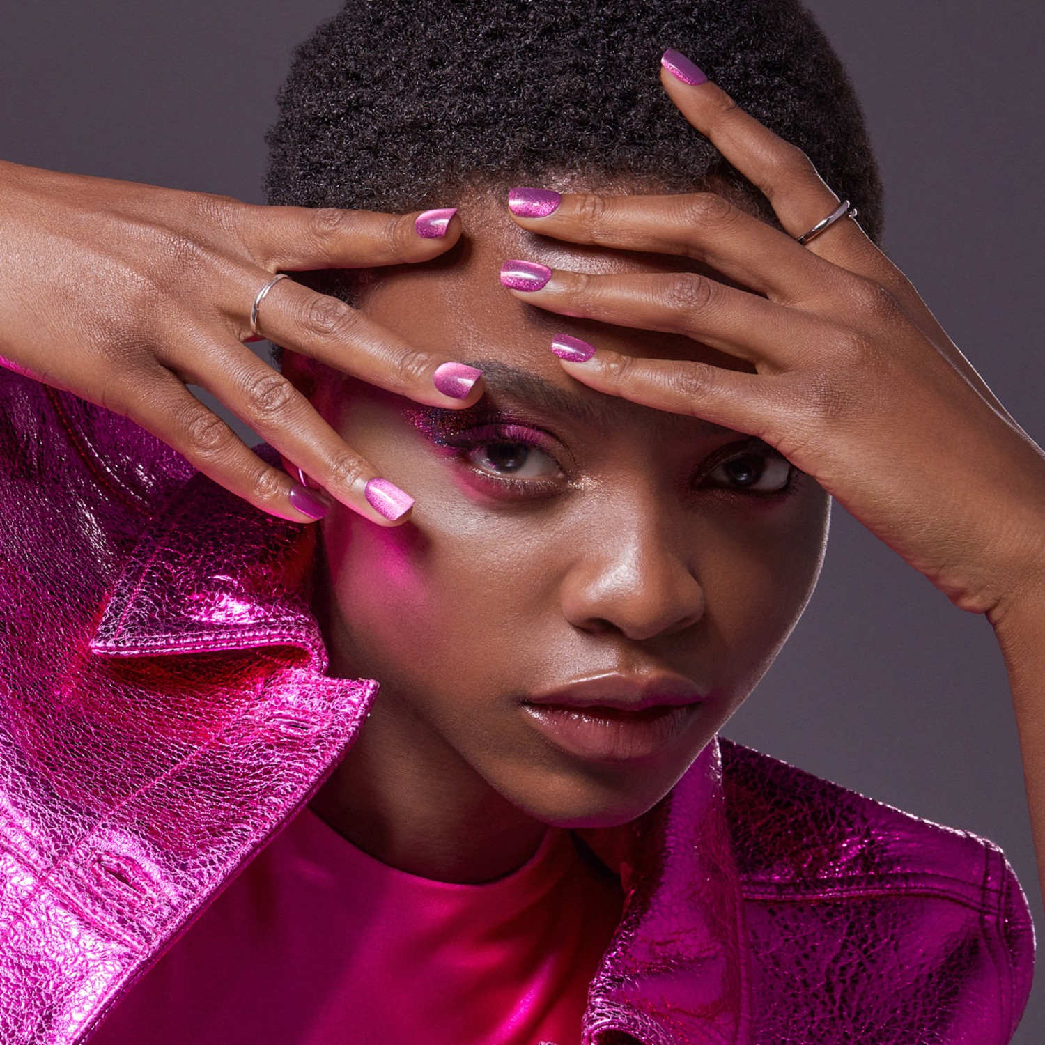 The image features a person with short hair, wearing a shiny, metallic pink jacket and a matching pink top. The individual has their hands placed on their forehead, partially covering their face. Their nails are painted in a glossy pink color, complementing the outfit. The background is a neutral gray, which helps to highlight the vibrant pink tones of the clothing and nails. The overall composition of the image is stylish and modern, with a focus on the bold fashion choices.