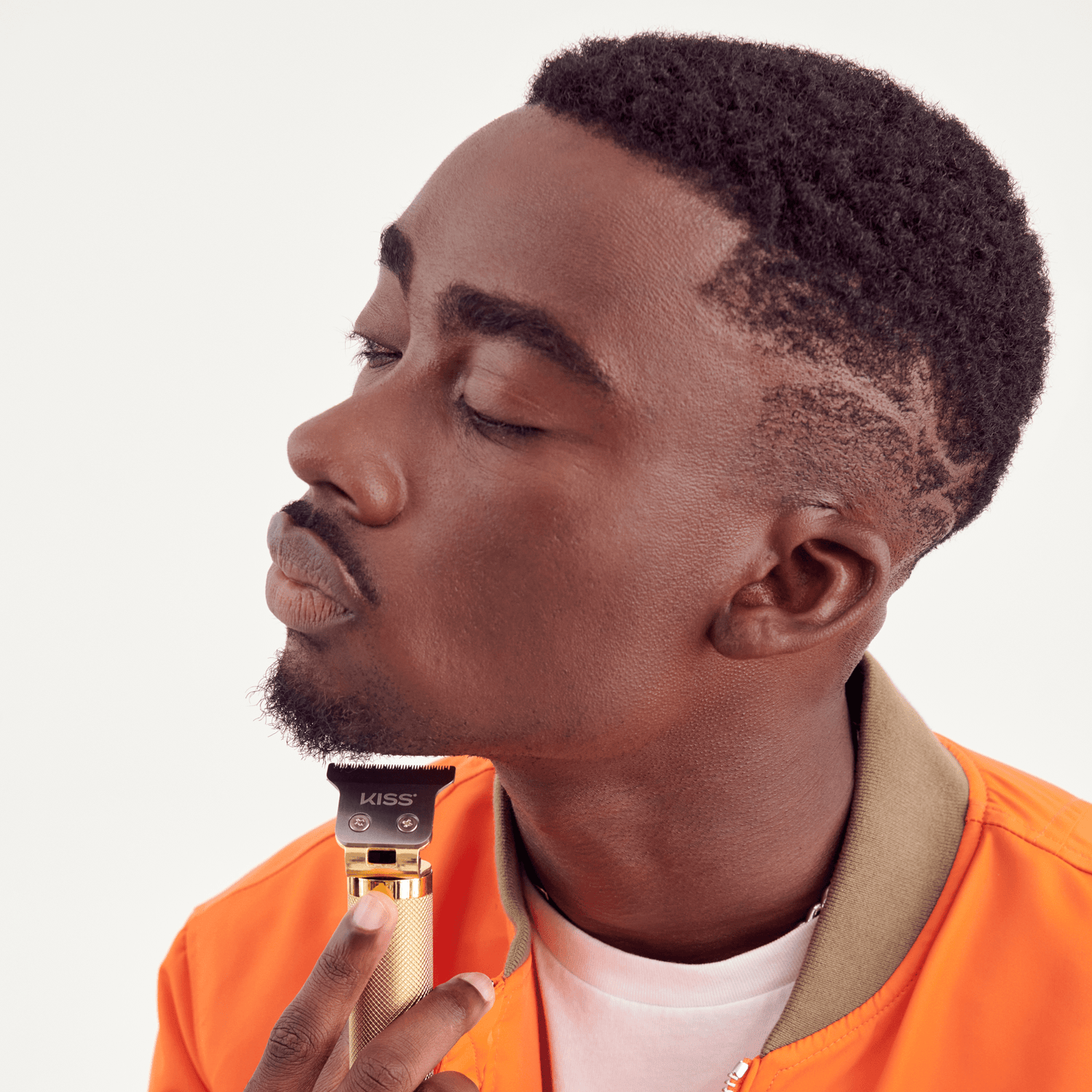 Man with short patterned hair wearing an orange jacket uses a gold and black KISS branded cordless trimmer to trim his beard along his jawline.