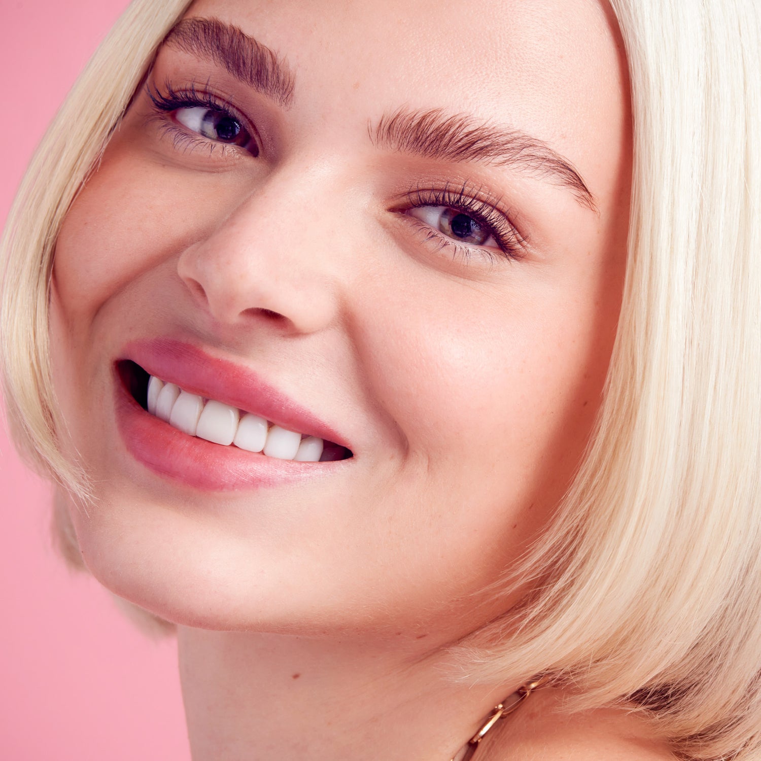 Close-up of a smiling woman with short blonde hair wearing natural, voluminous false eyelashes against a pink background.