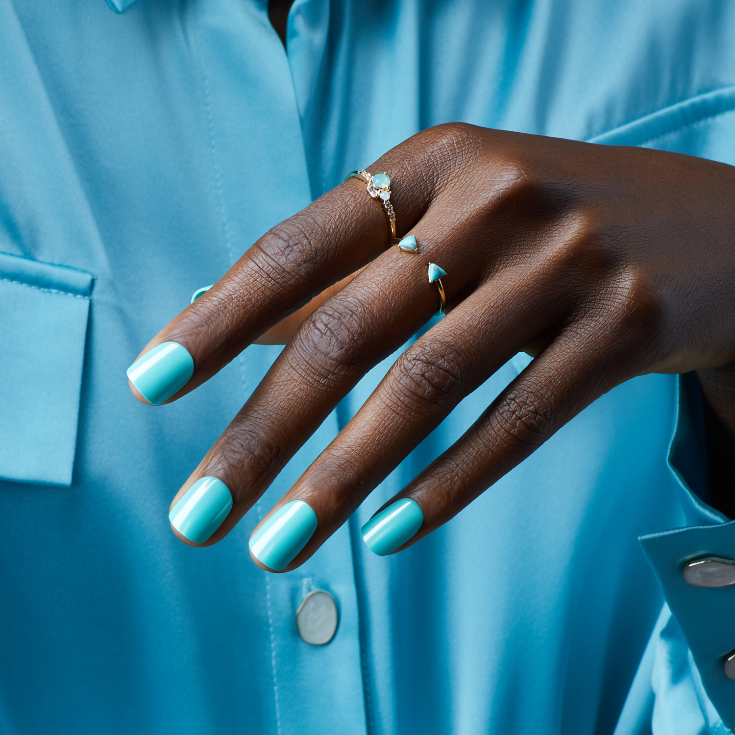 Dark-skinned hand wearing glossy light blue press-on nails, adorned with two delicate gold rings with turquoise stones, against a matching blue shirt.