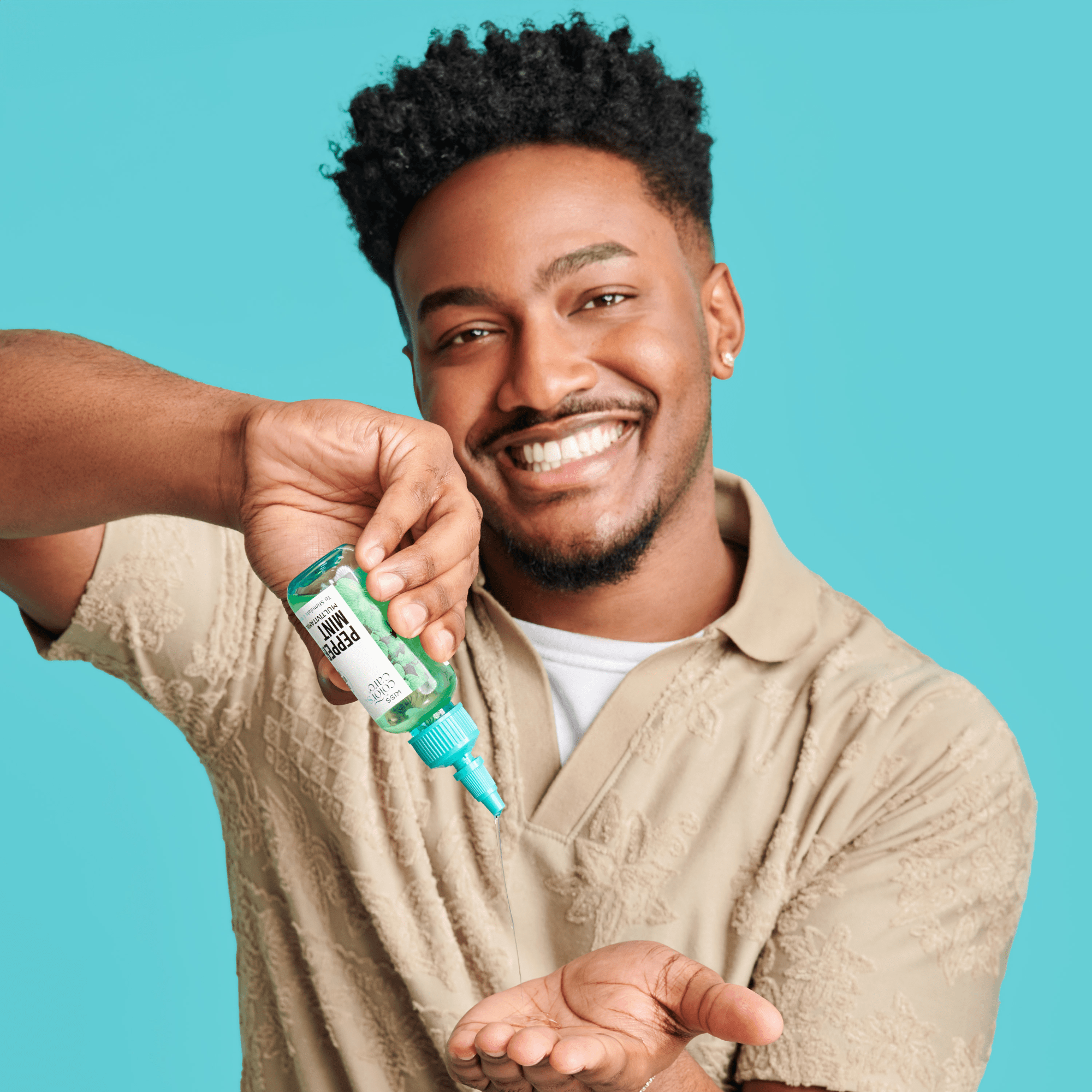 Smiling man with curly hair wearing a beige shirt pouring clear KISS Colors &amp; Care Multivitamin Oil - Peppermint from a green bottle onto his hand.