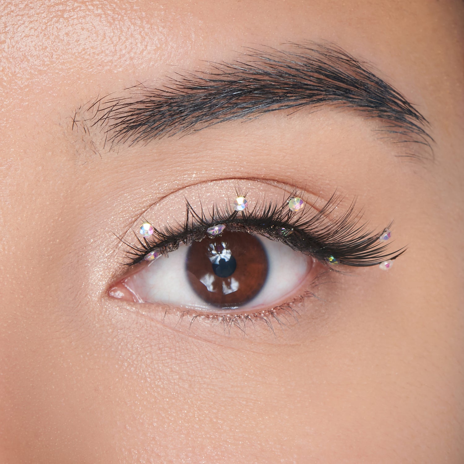 The image features a close-up of a human eye with a striking appearance. The eye has a rich brown iris and is framed by long, dark eyelashes. Delicate, sparkling embellishments are placed along the eyelid, adding a touch of glamour. The skin around the eye is smooth and has a subtle glow, enhancing the overall aesthetic. The focus on the eye creates an intimate and captivating look.