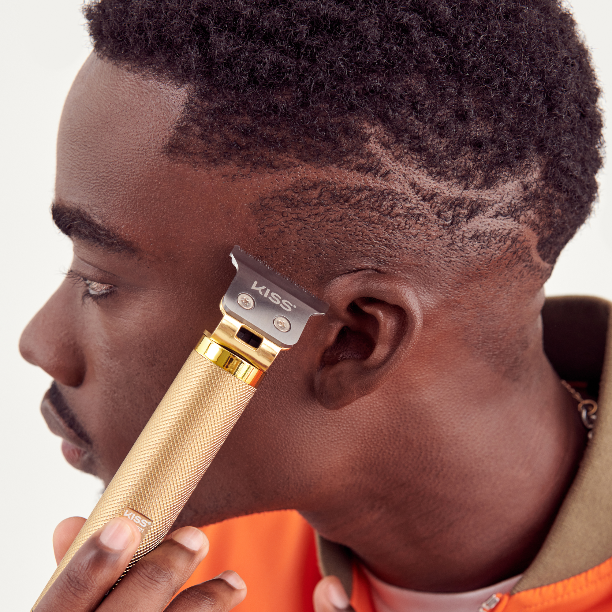 Man using a gold-colored KISS Precision Blade Cordless Trimmer near his hairline, showing detailed hair design on the side of his head.
