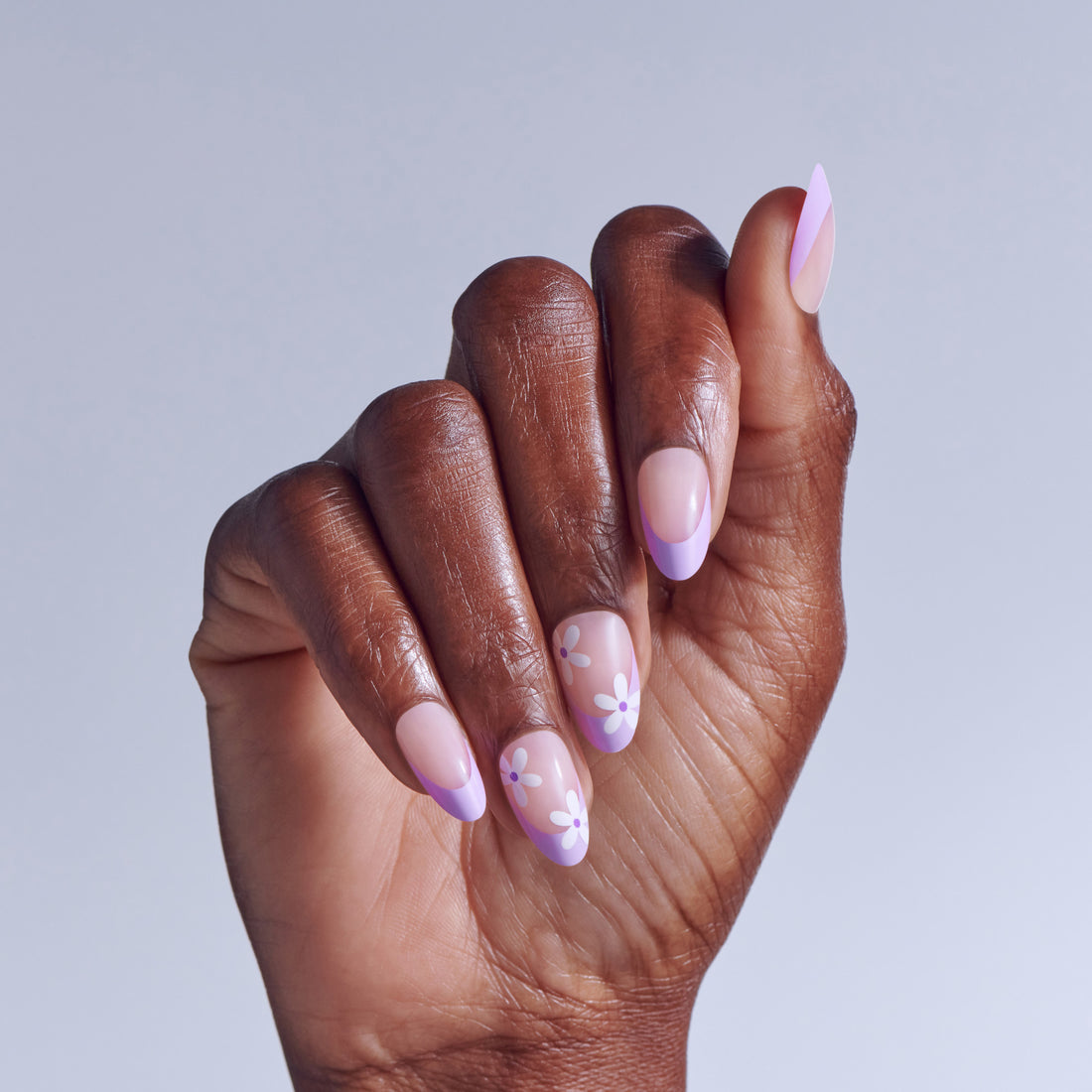 Hand with oval nails featuring pink base and purple floral designs against light blue background.
