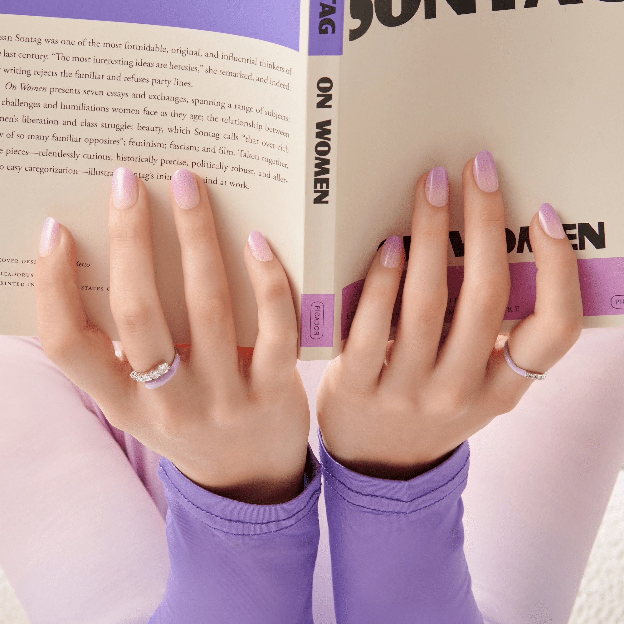 The image features a pair of hands holding an open book titled "On Women." The hands are elegantly manicured with a soft lavender nail polish. One hand is adorned with a delicate ring, while the other has a simple band. The background appears to be a soft pastel color, and the person is wearing a matching lavender top. The overall aesthetic is calm and stylish, emphasizing a moment of reading and reflection.