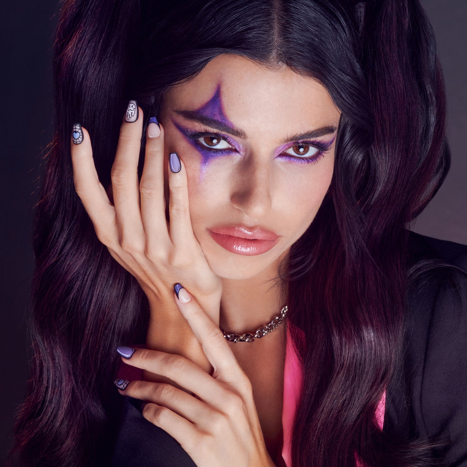 Woman with long dark hair styled in pigtails wearing Halloween-inspired purple eye makeup and light volume false eyelashes, showing detailed nail art on lavender and neutral nails.