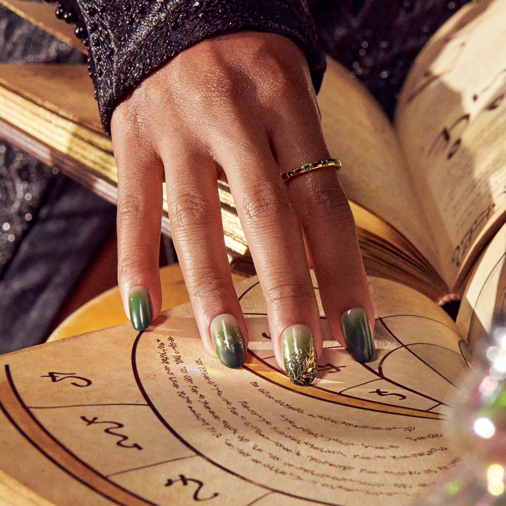 Hand with green ombré press-on nails featuring gold foil accents, resting on an open ancient book with astrological symbols.