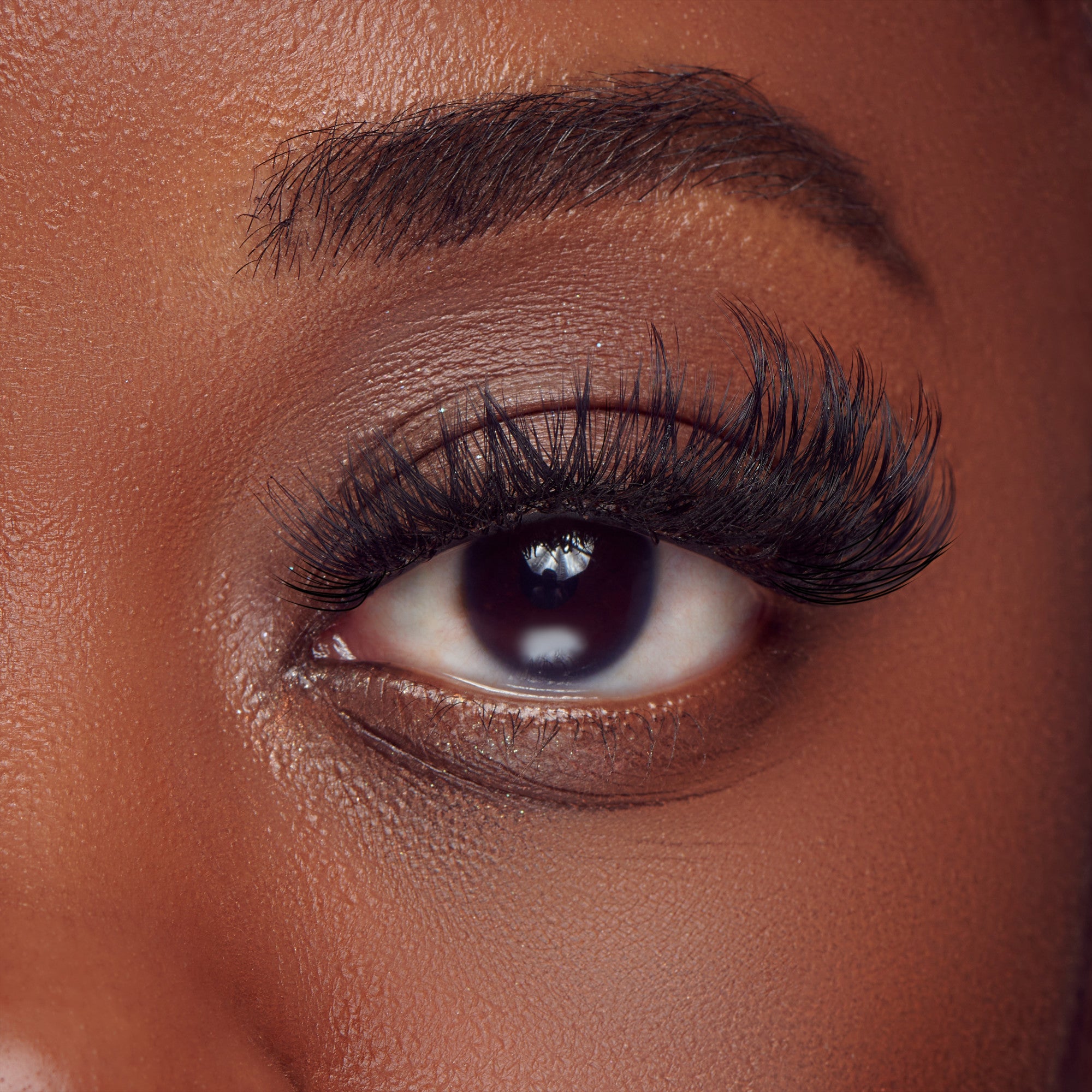 The image features a close-up of an eye, showcasing long, dramatic eyelashes. The skin around the eye is smooth and has a warm tone, highlighting the natural beauty of the individual. The eyebrow is well-groomed and arches gracefully above the eye. The iris is dark, and the overall composition emphasizes the eye&