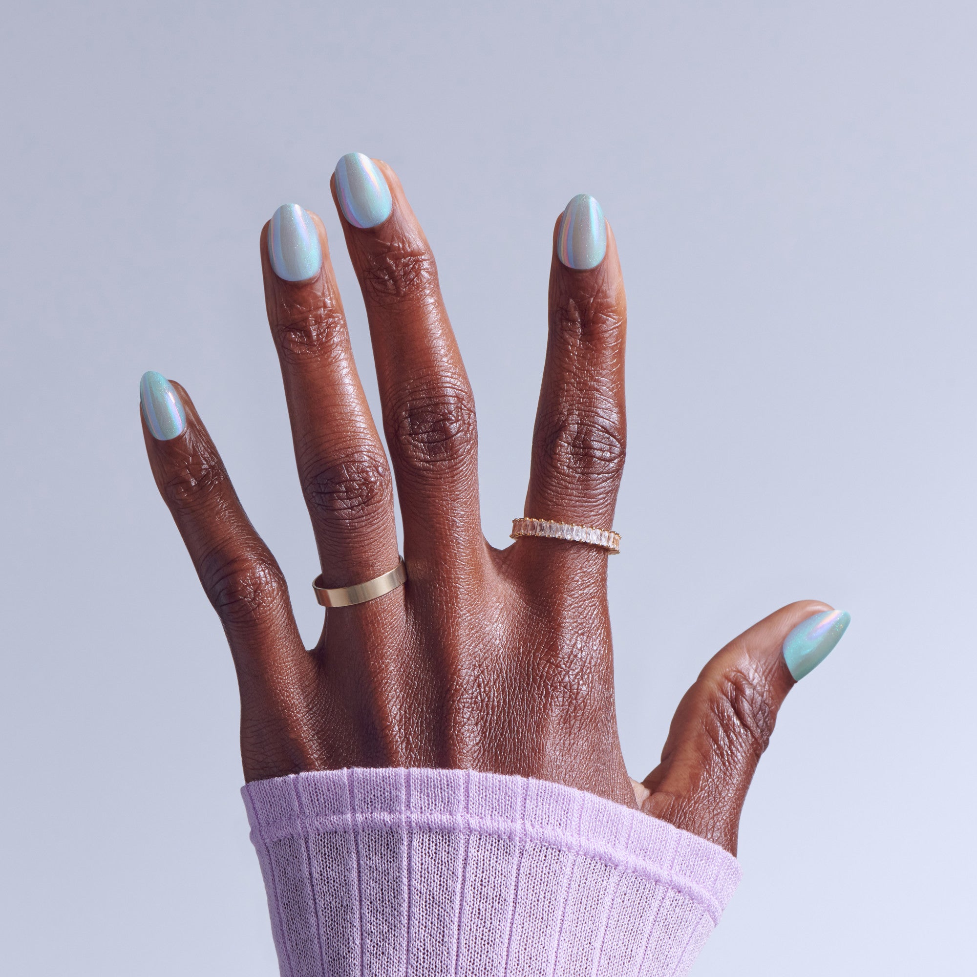 Hand with iridescent nail polish manicure and silver rings against lavender background.